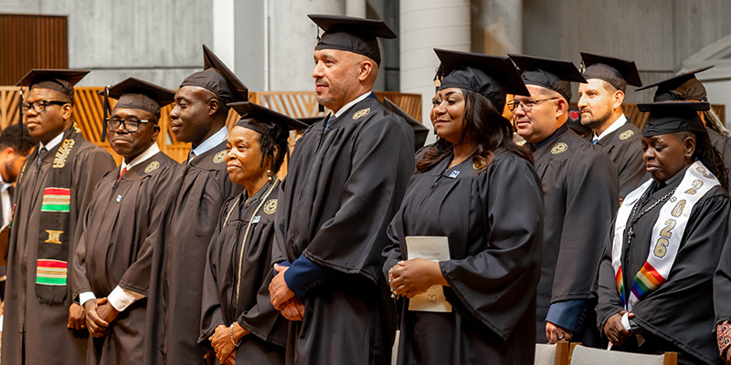 Graduates in caps and gowns stand in a row during a ceremony, facing forward in a chapel.