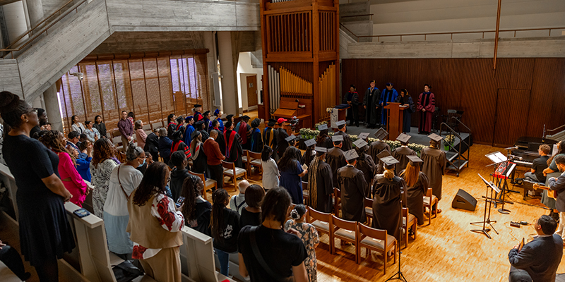 A graduation ceremony in a modern chapel, with rows of graduates in caps and gowns facing a stage where speakers stand at a podium, while attendees watch from seats and along the sides.