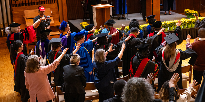 Group of people in academic gowns stand with hands raised in prayer during a ceremony in a chapel.