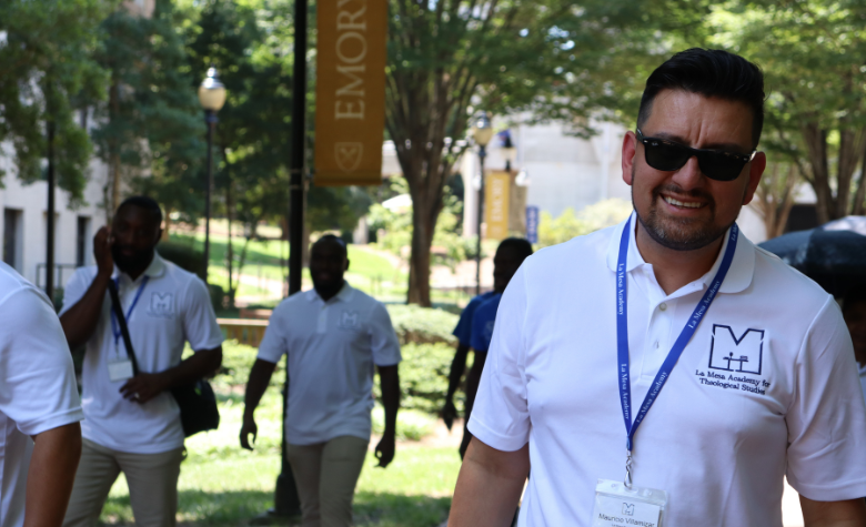 Person wearing a white La Mesa polo shirt and conference badge walks outdoors on a campus pathway, with several other attendees and Emory banners visible in the background.
